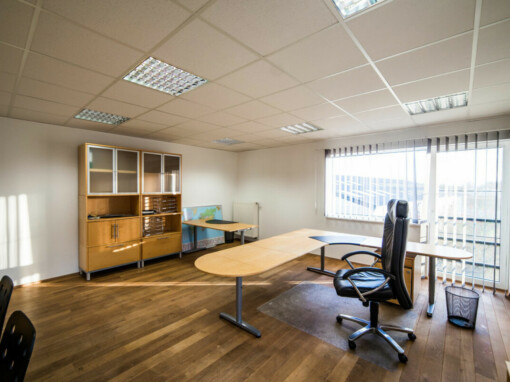 Un bureau professionnel avec du parquet au sol, un armoire en bois, un grand bureau en L et une chaise de bureau, des vitres qui donnent sur l'extérieur